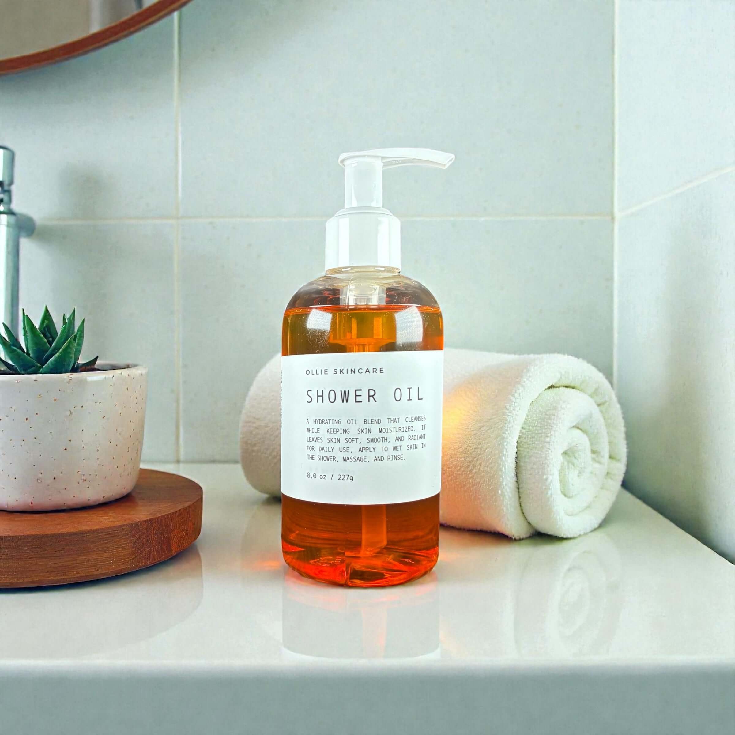 Ollie Skincare Shower Oil bottle on a bathroom counter with a towel and plant.
