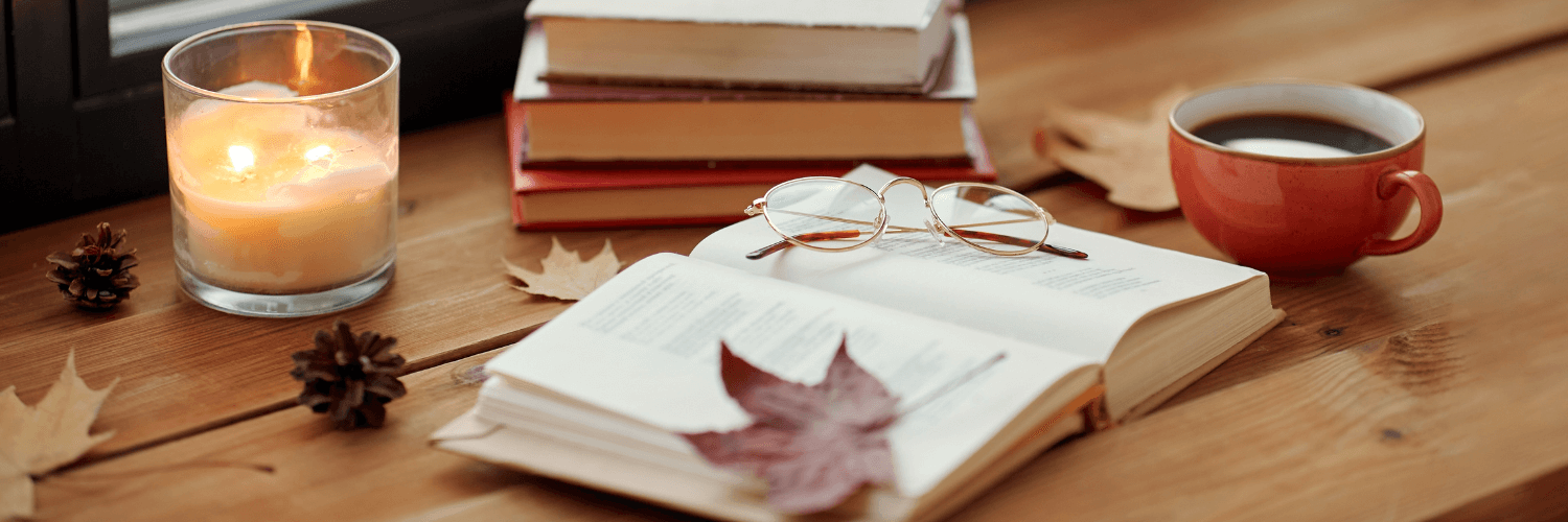 cozy autumn scene with a candle, open book, glasses, coffee cup, and fallen leaves, perfect for the Glowup Challenge: Fall Edition.