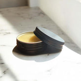 Calendula Herbal Salve in a dark tin container with lid open, revealing creamy balm inside on marble surface.