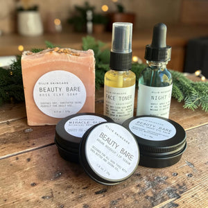 Glow Ritual gift set products: rose clay facial soap, face toner mist, night oil and three balm tins on a wooden table.
