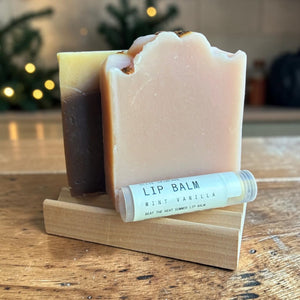 Two goat milk soap bars on a wooden soap tray with a Mint Vanilla lip balm, from The Soap Lover gift set.