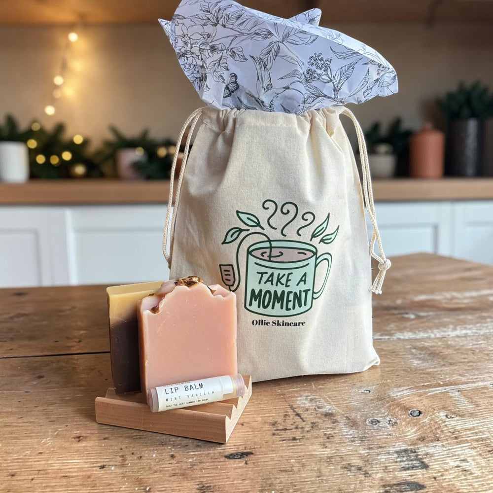 The Soap Lover gift set: two goat milk soap bars on a wooden soap tray with a lip balm, beside a 'Take a Moment' cotton gift bag.