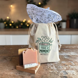 The Soap Lover gift set: two goat milk soap bars on a wooden soap tray with a lip balm, beside a 'Take a Moment' cotton gift bag.