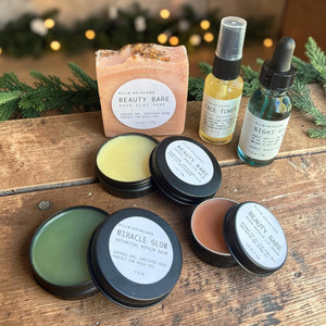 Glow Ritual gift set with open balm tins, rose clay facial soap, face toner mist and night oil on a wooden board.