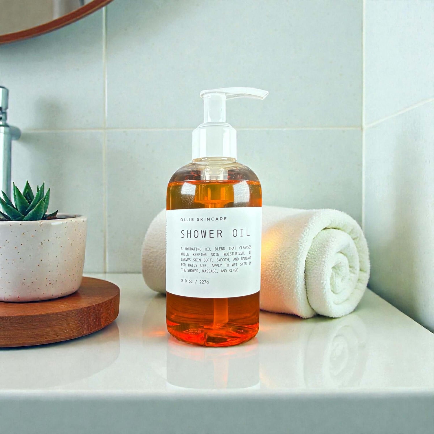 Ollie Skincare Shower Oil bottle on a bathroom counter with a towel and plant.