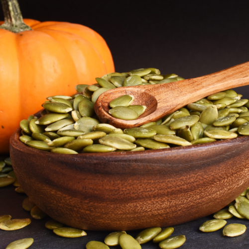 Pumpkin seeds in a wooden bowl with a wooden spoon, next to a pumpkin on a dark background.
