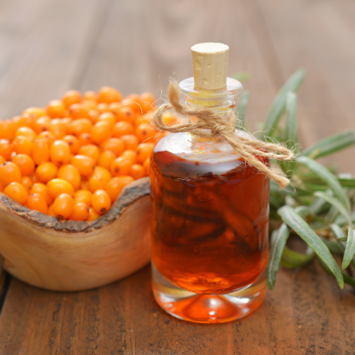 Bottle of amber-colored liquid with a wooden cap, surrounded by sea buckthorn berries and green leaves on a wooden surface.